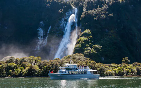 Small boat cruising near waterfall in Milford Sound, New Zealand.