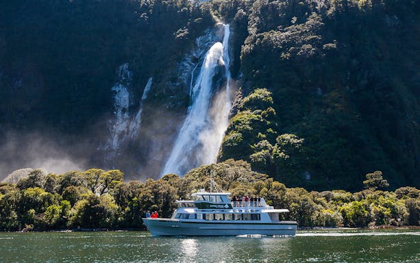 Small boat cruising near waterfall in Milford Sound, New Zealand.