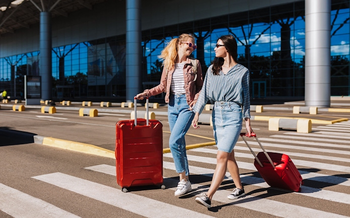 Two women with luggage crossing a taxi lane at an airport.