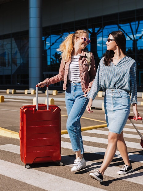 Two women with luggage crossing a taxi lane at an airport.