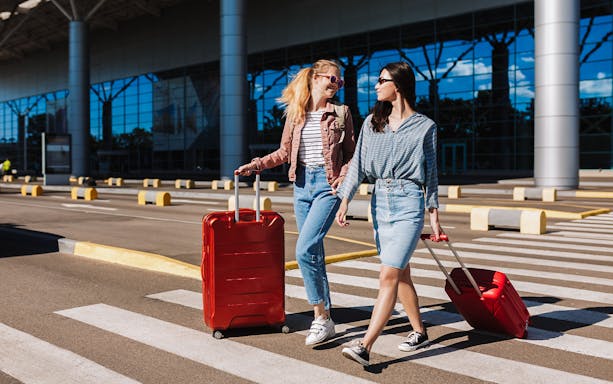 Two women with luggage crossing a taxi lane at an airport.