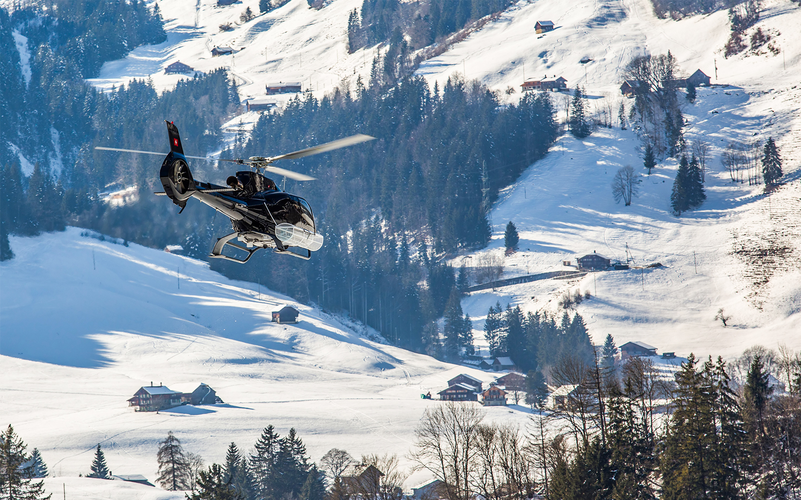 Ein Helikopter fliegt über eine Schneelandschaft mit Tannen und kleinen Häusern