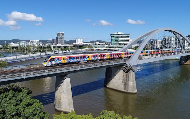 Train crossing bridge in Brisbane with city skyline in background.