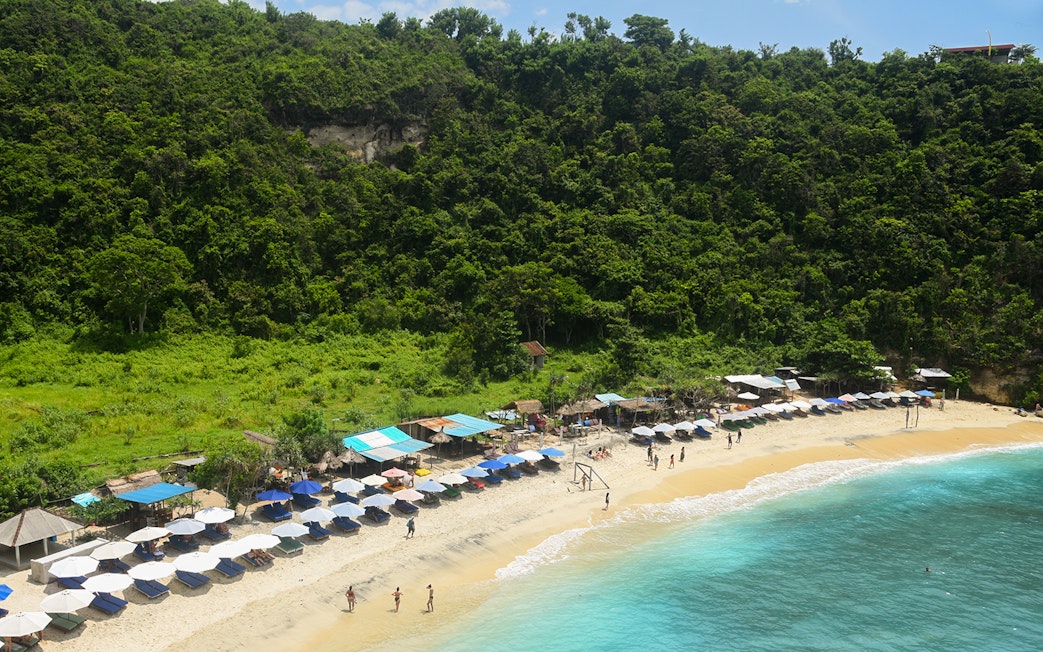 Beach with umbrellas and tourists at Nusa Penida Island, Indonesia, surrounded by lush greenery.