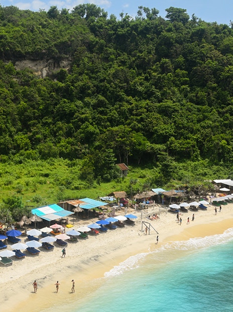 Beach with umbrellas and tourists at Nusa Penida Island, Indonesia, surrounded by lush greenery.