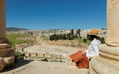Woman in hat sitting at Jerash ruins overlooking ancient colonnade and cityscape.