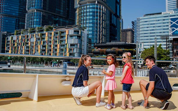 Kids with Captain Cook Sydney Harbour Cruise staff on deck with city skyline.