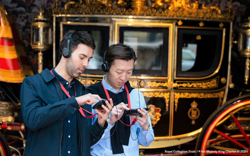 Visitors using audio guides at Buckingham Palace with a royal carriage in the background.