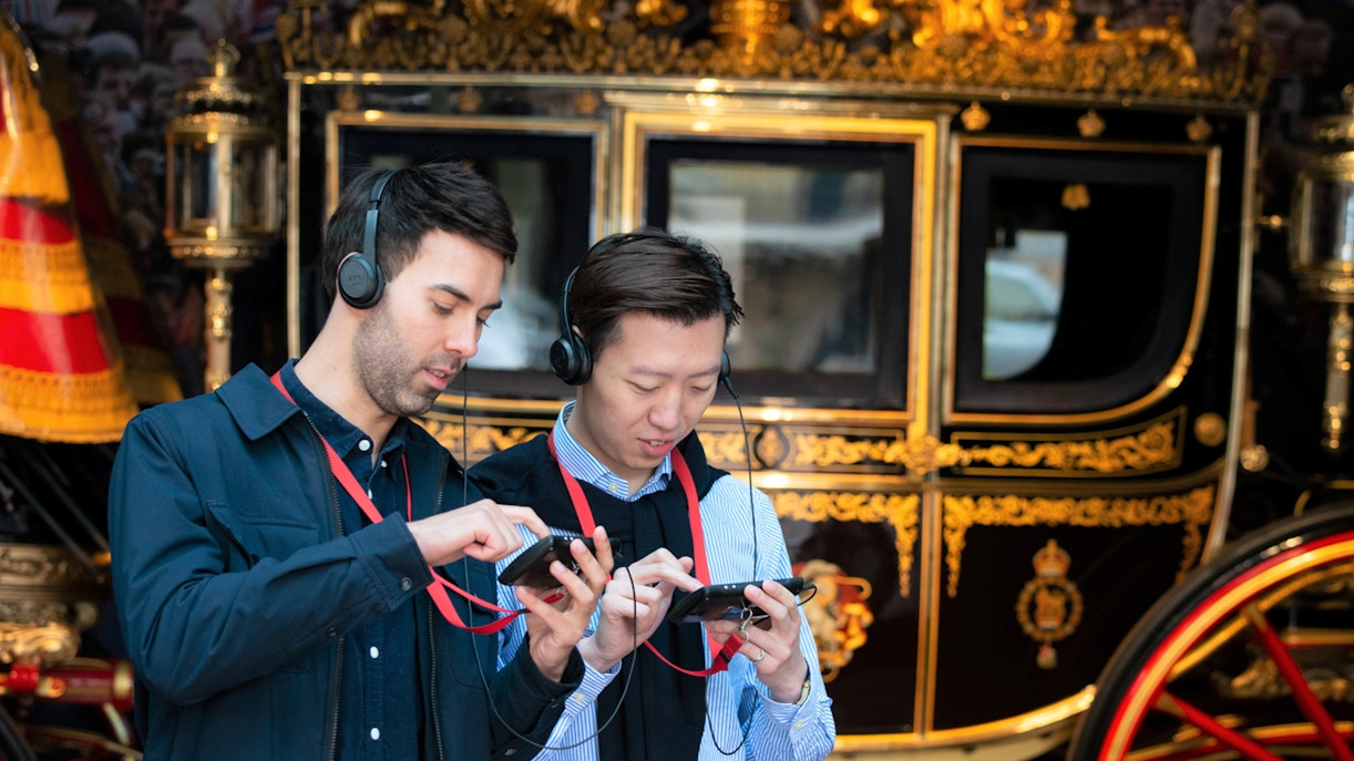 Visitors using audio guides at Buckingham Palace with a royal carriage in the background.