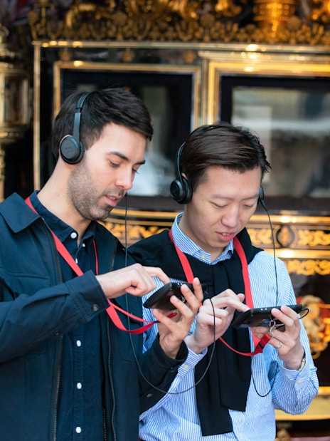 Visitors using audio guides at Buckingham Palace with a royal carriage in the background.