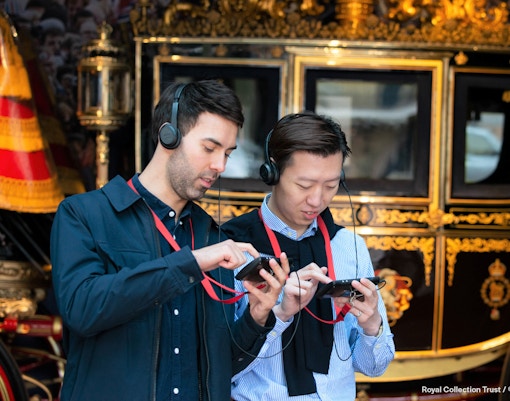 Visitors using audio guides at Buckingham Palace with a royal carriage in the background.