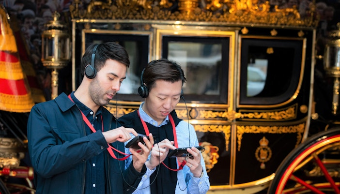 Visitors using audio guides at Buckingham Palace with a royal carriage in the background.