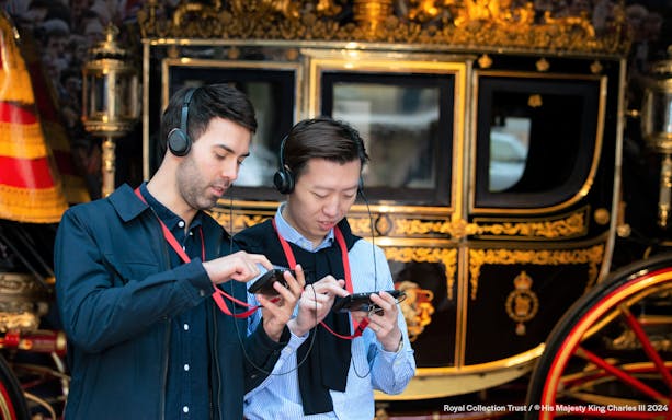 Visitors using audio guides at Buckingham Palace with a royal carriage in the background.