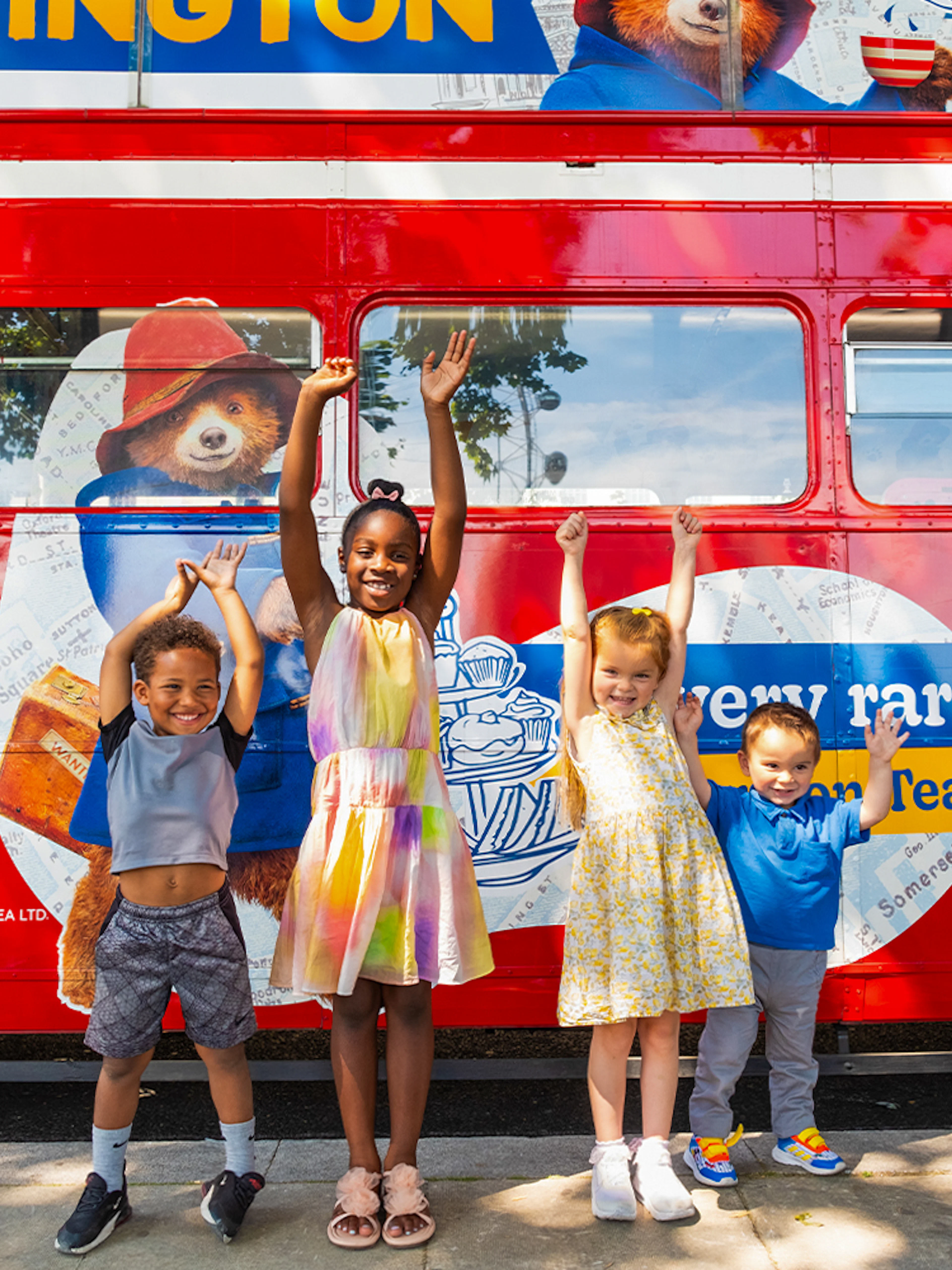 Children smiling in front of the Paddington Afternoon Tea Bus in London.