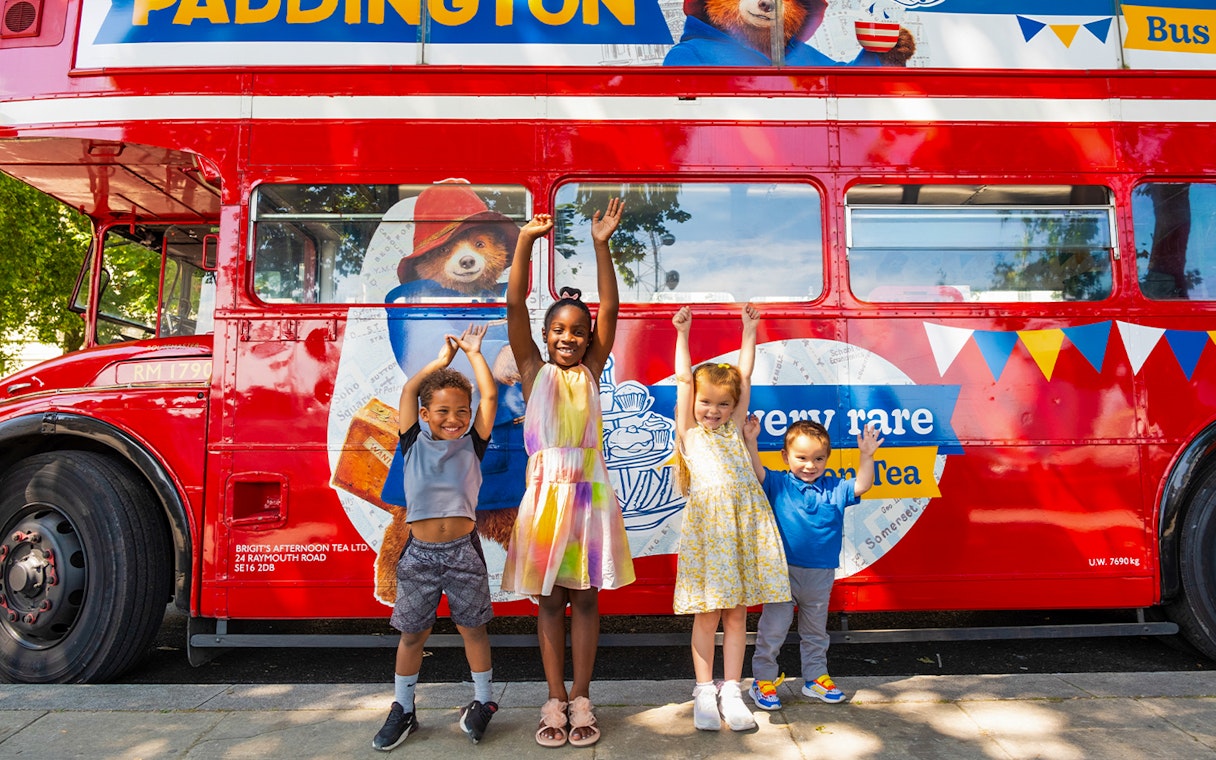 Children smiling in front of the Paddington Afternoon Tea Bus in London.