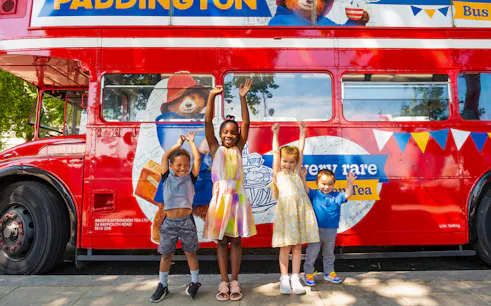 Children smiling in front of the Paddington Afternoon Tea Bus in London.