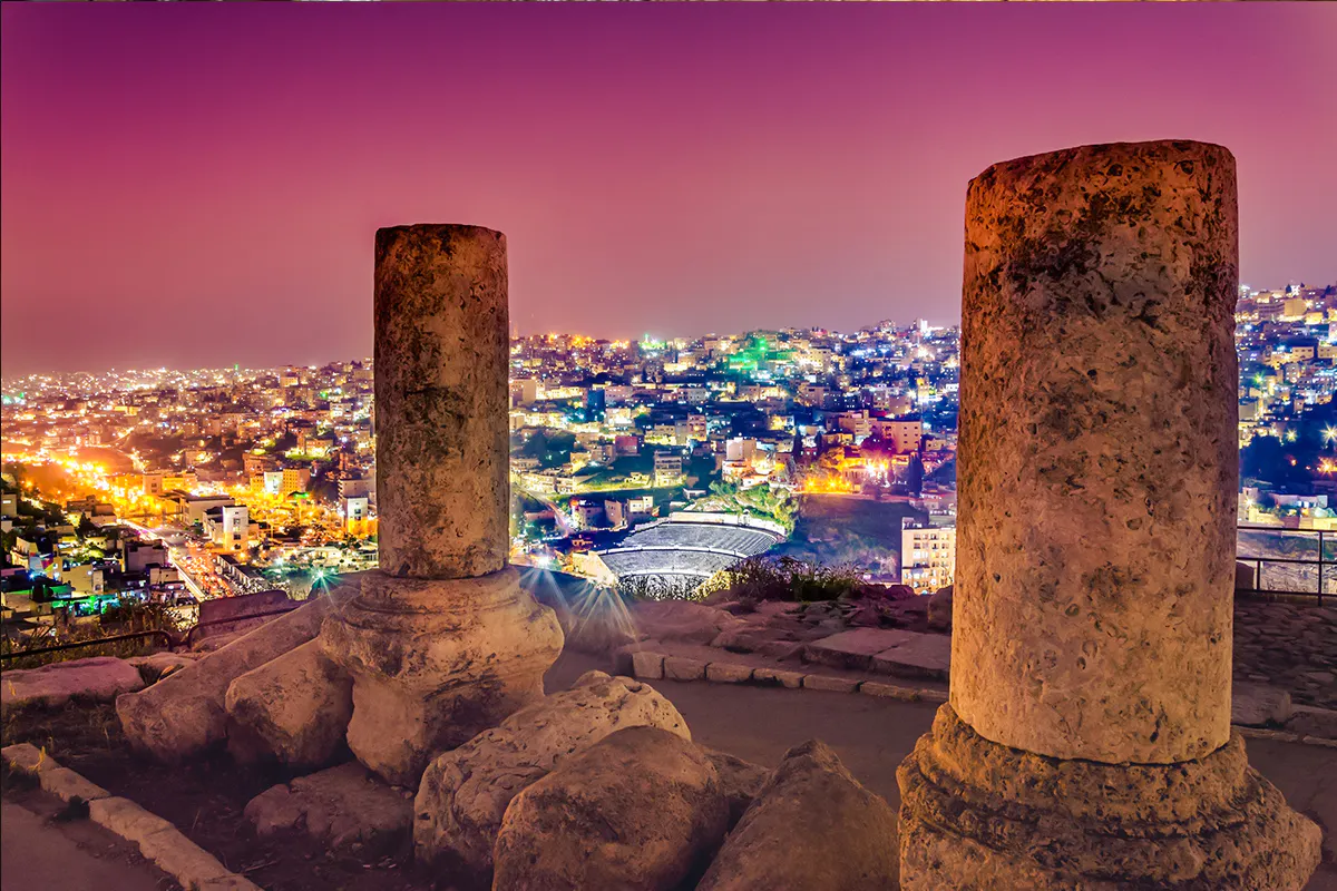Ancient columns overlooking Amman cityscape and Roman Amphitheater at night.