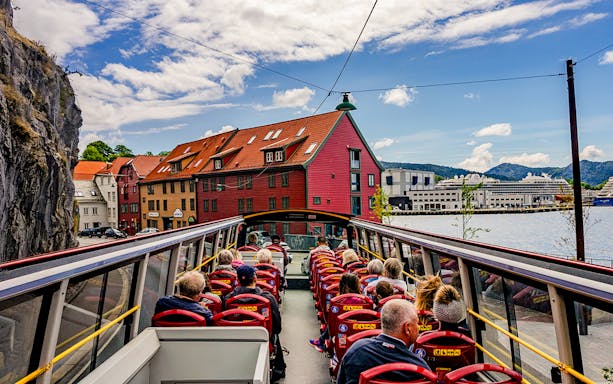 Bergen Hop on Hop off bus with tourists passing colorful waterfront buildings.