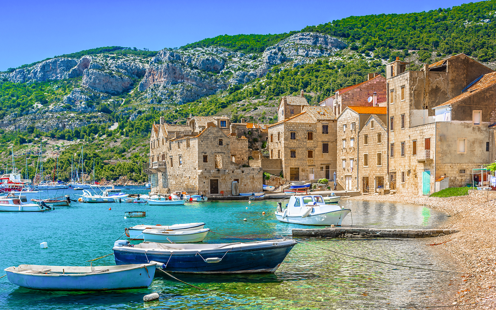 Boats docked by stone buildings on Vis Island, Croatia, with rocky hills in the background.