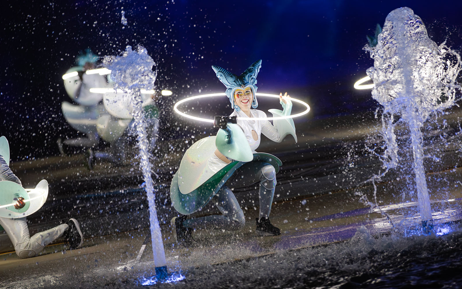 Performer with glowing hoop at Kiss of the Sea Show, surrounded by water fountains.