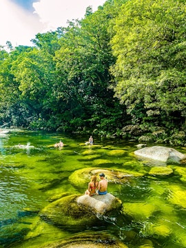 People swimming and sitting on rocks in a river surrounded by lush greenery in Daintree National Park.