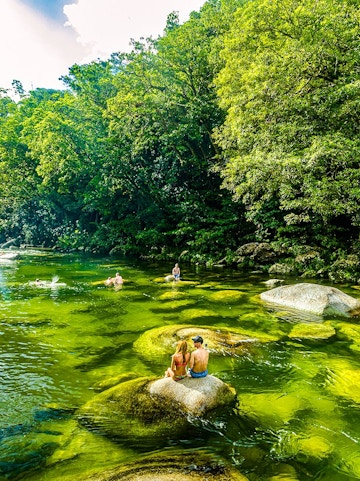 People swimming and sitting on rocks in a river surrounded by lush greenery in Daintree National Park.