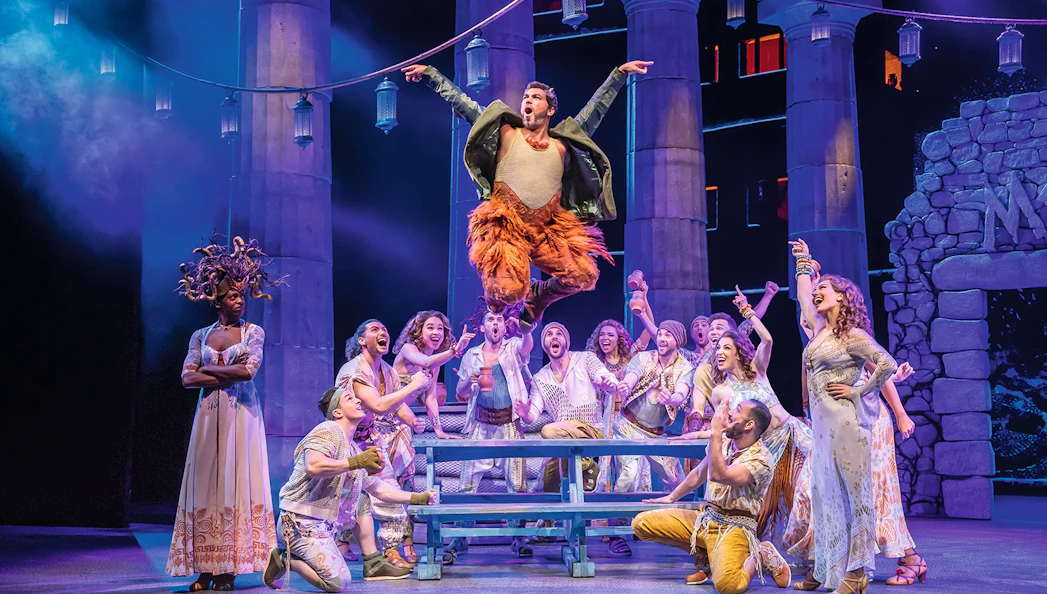 Stage performance of Disney's Hercules with actors in ancient Greek costumes, featuring a lively scene.