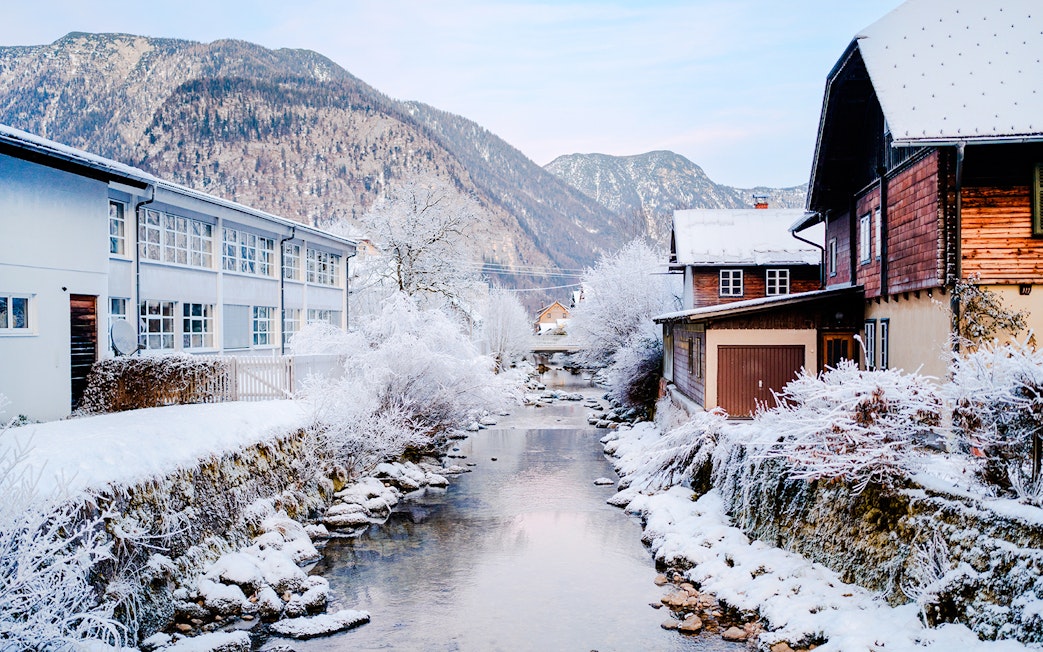 Snow-covered stream and buildings in Hallstatt old town, Austria, with mountains in the background.