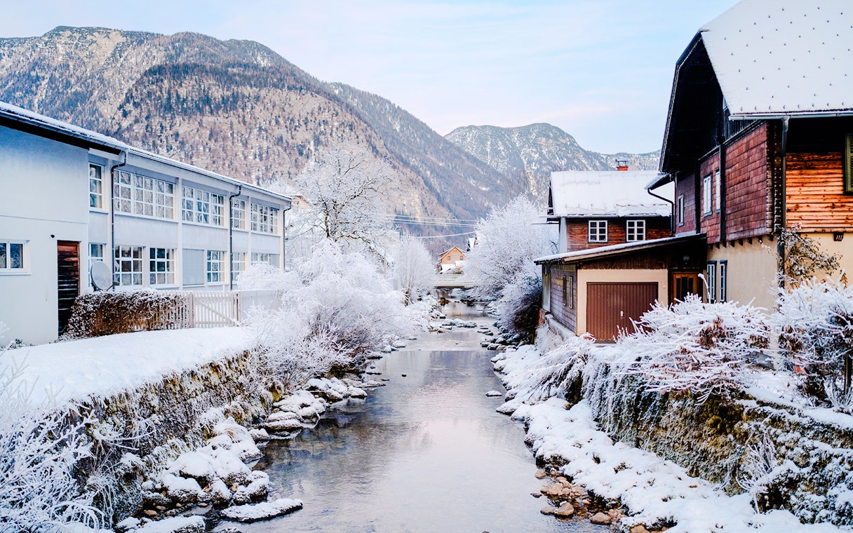 Snow-covered stream and buildings in Hallstatt old town, Austria, with mountains in the background.
