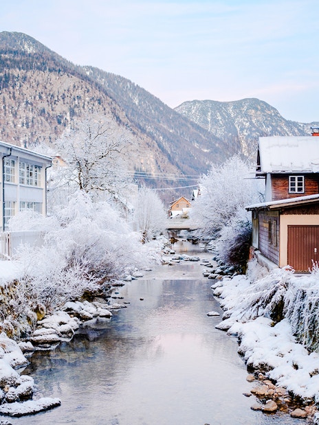 Snow-covered stream and buildings in Hallstatt old town, Austria, with mountains in the background.