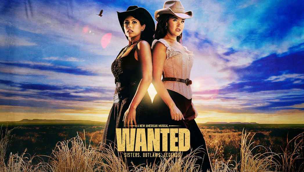 Two women in cowboy hats stand back-to-back in a field under a sunset sky, promoting "Wanted" musical on Broadway.
