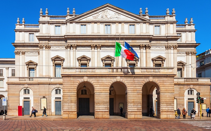 La Scala opera house facade in Milan with Italian and EU flags.