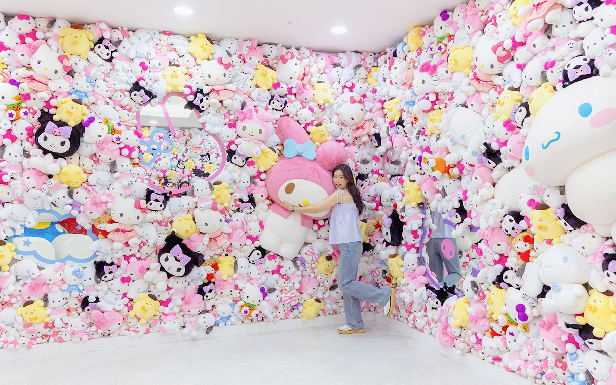 Tourist hugging large plush at Hello Kitty Island surrounded by colorful character toys.