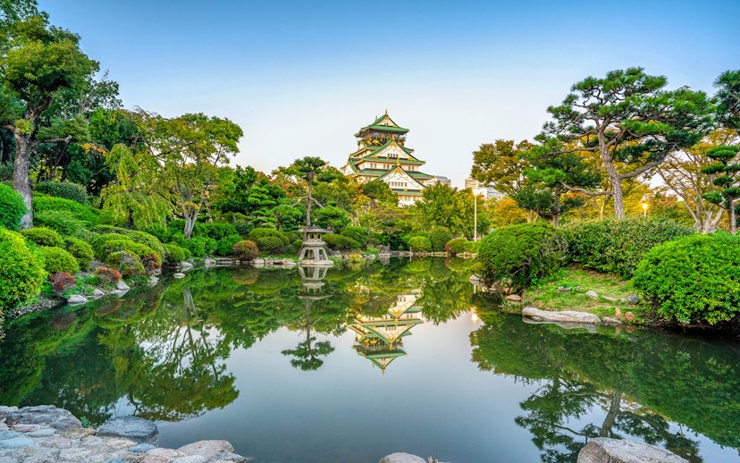 Osaka Castle reflected in a serene garden pond, surrounded by lush greenery.