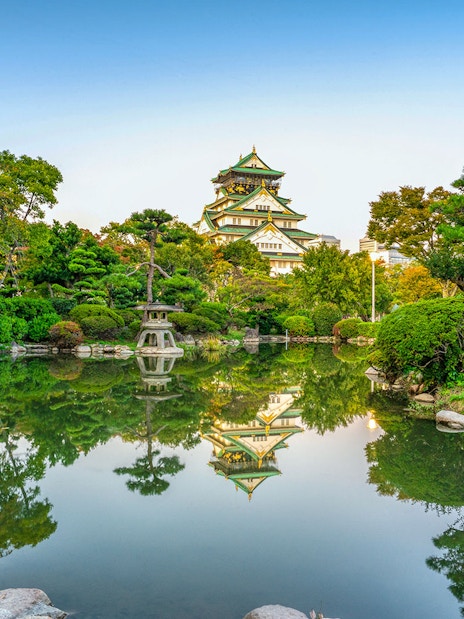 Osaka Castle reflected in a serene garden pond, surrounded by lush greenery.