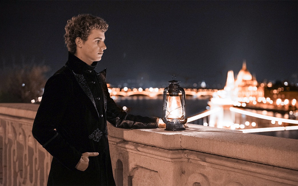 Person in dark attire holding lantern on Buda Castle tour, Budapest, with city lights in background.