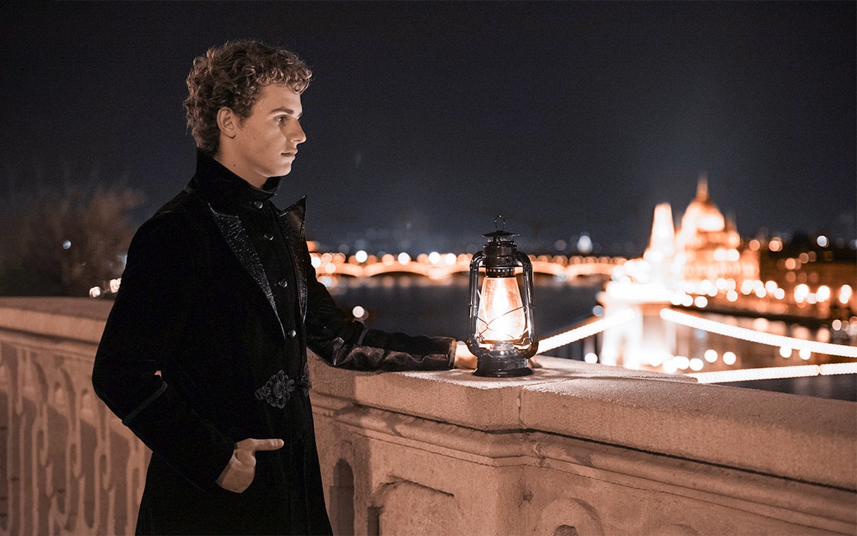 Person in dark attire holding lantern on Buda Castle tour, Budapest, with city lights in background.