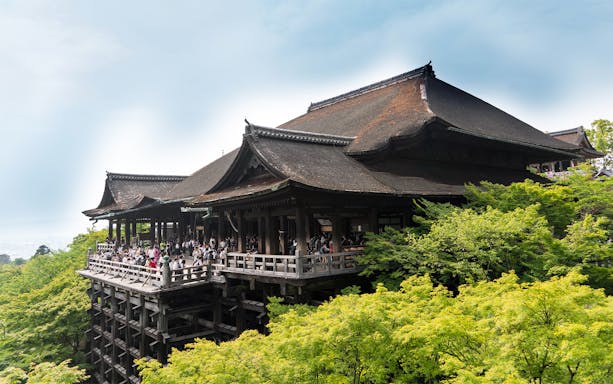 Kiyomizu-dera temple in Kyoto with visitors on the wooden terrace overlooking lush green forest.