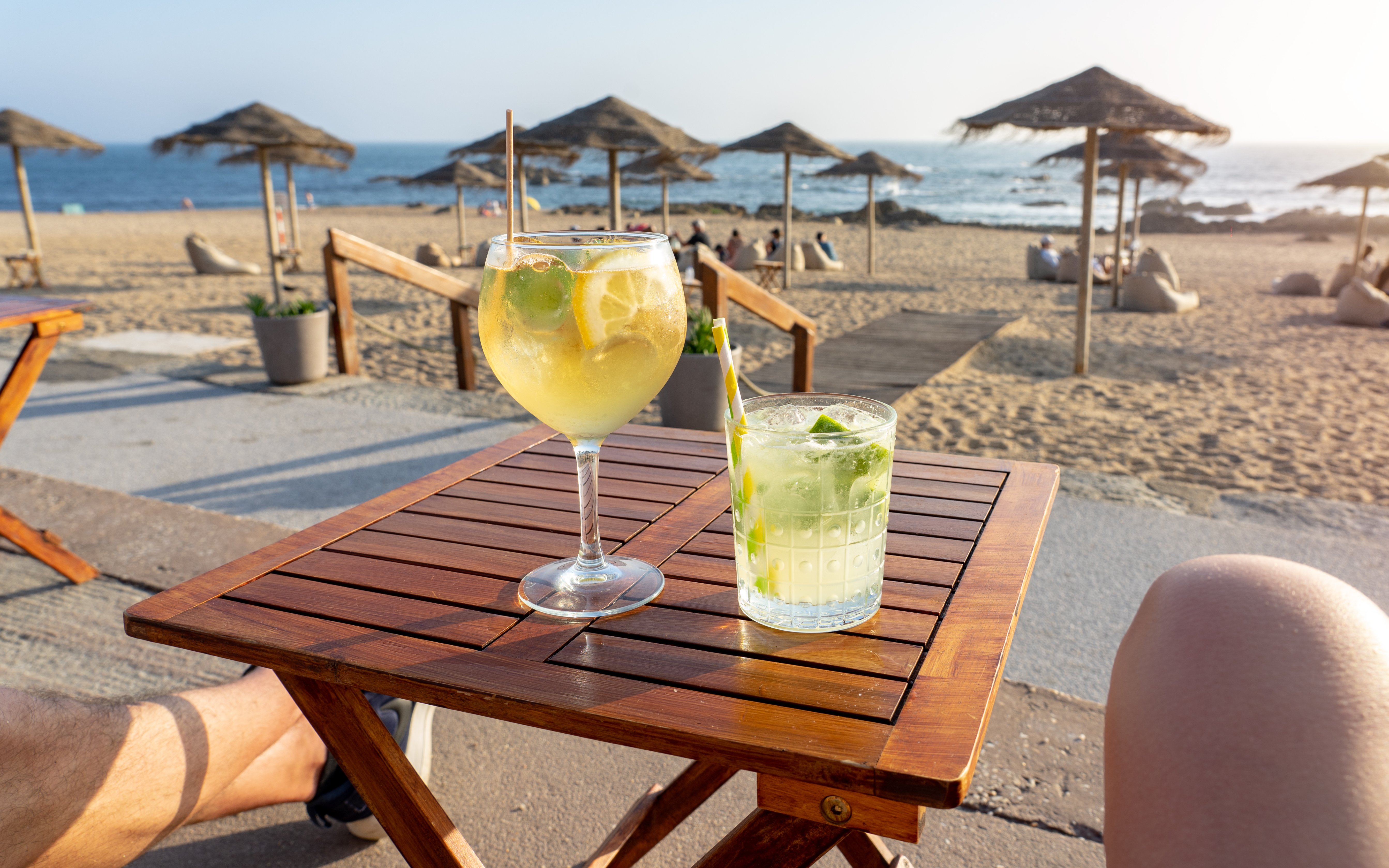 Drinks on a wooden table at a beach bar with ocean view and straw umbrellas.