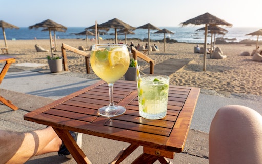 Drinks on a wooden table at a beach bar with ocean view and straw umbrellas.