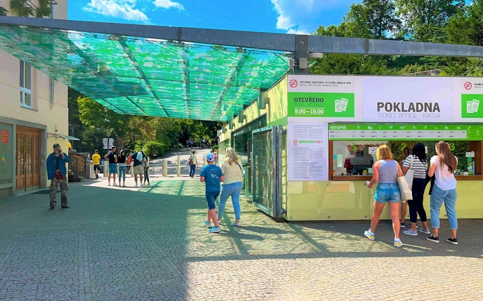 Visitors at Prague Zoo ticket office under a green canopy.