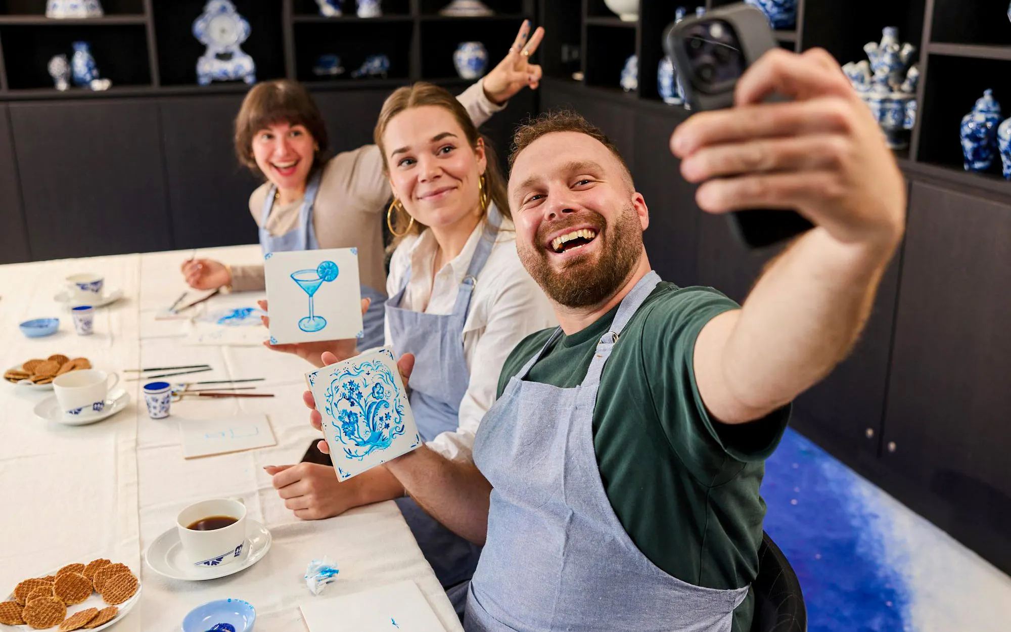 Guests painting tiles at a Dutch blue tile workshop, showing their artwork and taking a selfie.