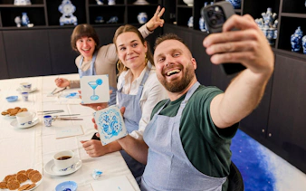 Guests painting tiles at a Dutch blue tile workshop, showing their artwork and taking a selfie.