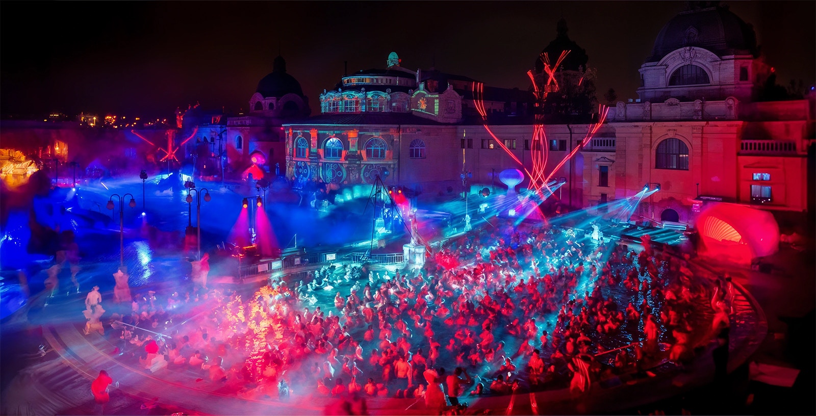Nighttime party at Szechenyi Bath in Budapest with colorful lights and a large crowd.