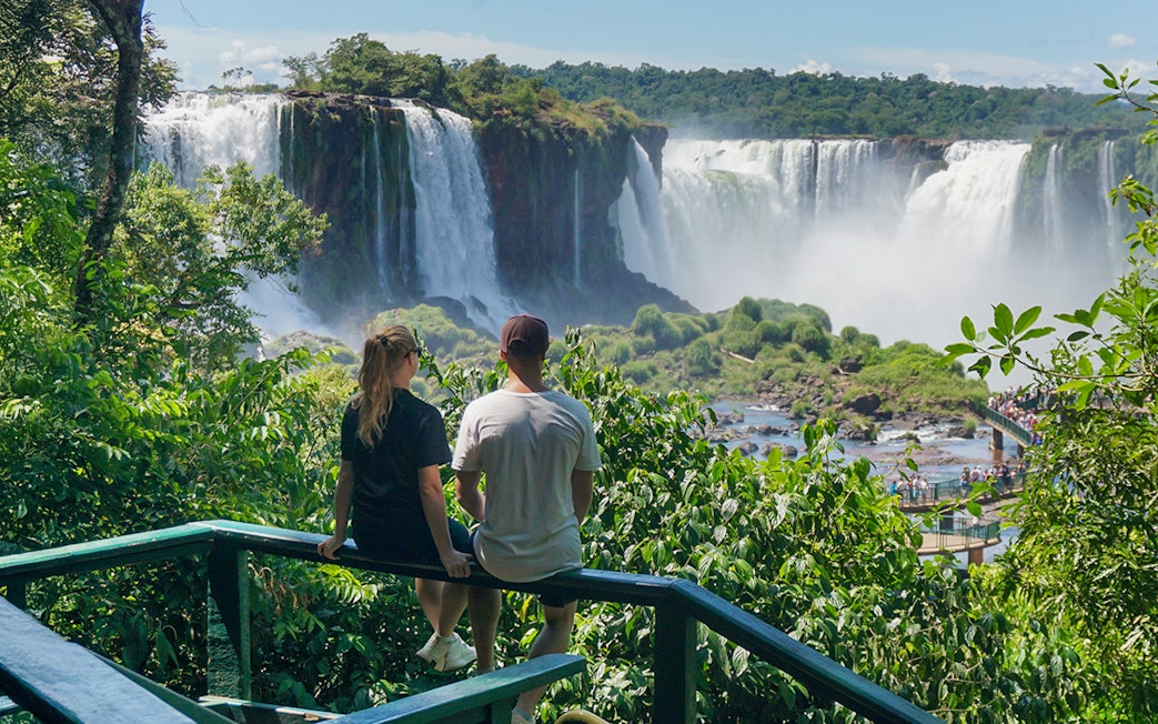 Man and woman viewing Iguazu Falls from a lush green lookout point.