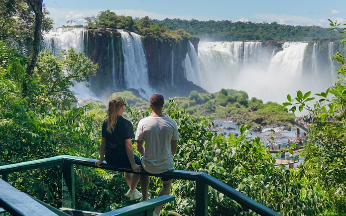 Man and woman viewing Iguazu Falls from a lush green lookout point.
