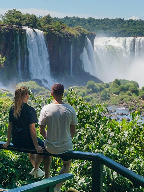 Man and woman viewing Iguazu Falls from a lush green lookout point.