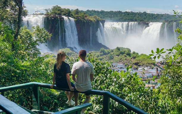 Man and woman viewing Iguazu Falls from a lush green lookout point.