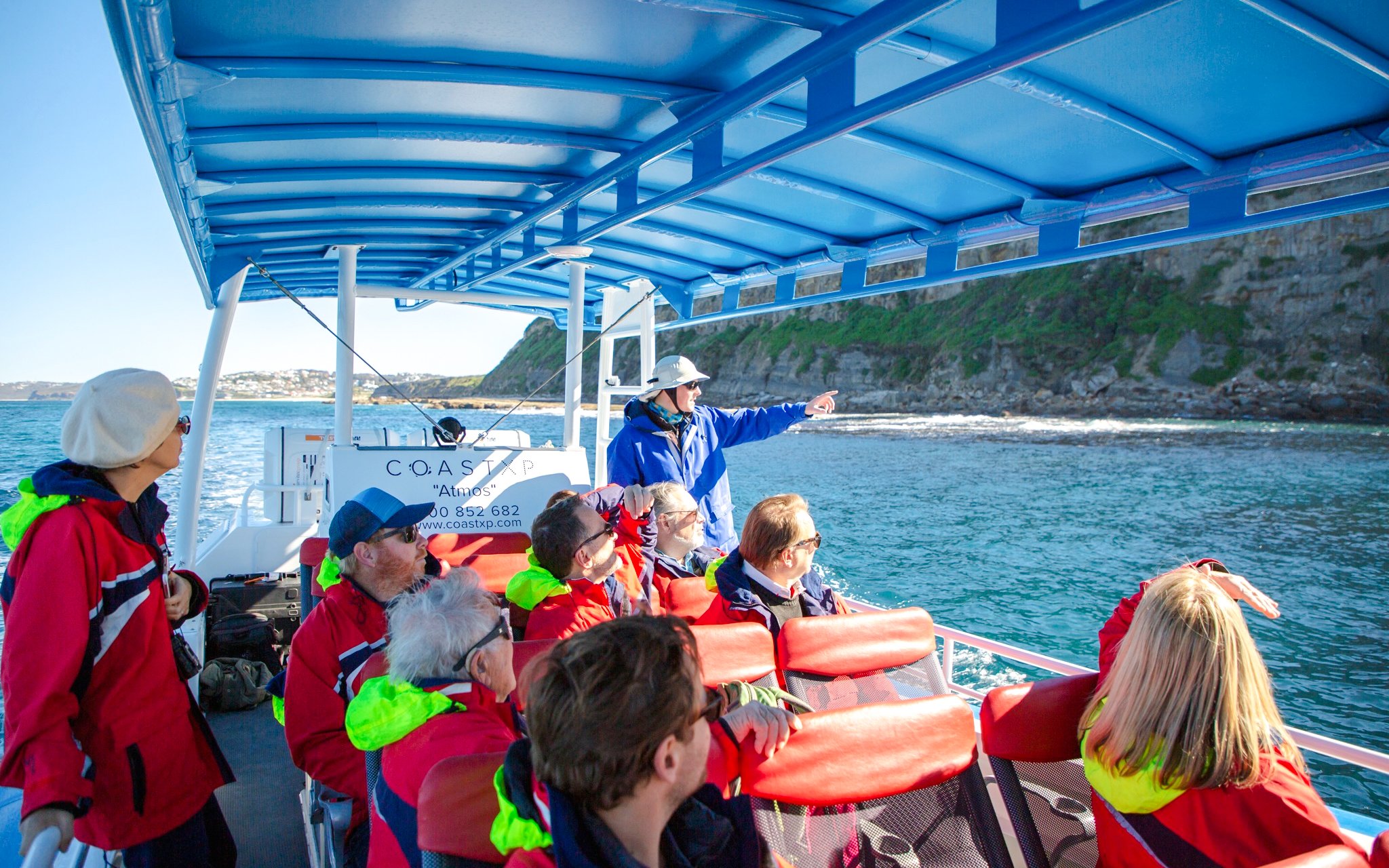 Tourists on a boat tour in Newcastle, guide pointing at coastal cliffs.