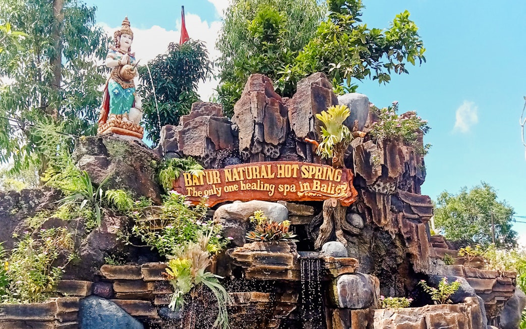 Batur Natural Hot Spring entrance sign with statue and lush greenery in Bali.